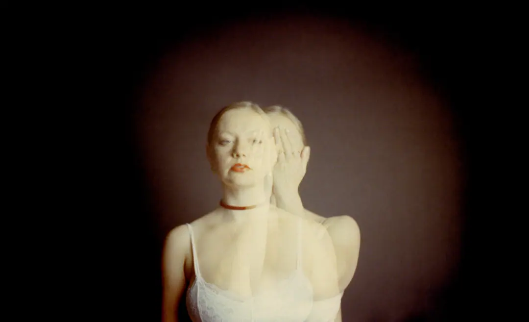 Woman in white lace top with translucent double exposure, one facing forward and one with hand covering face against dark background.
