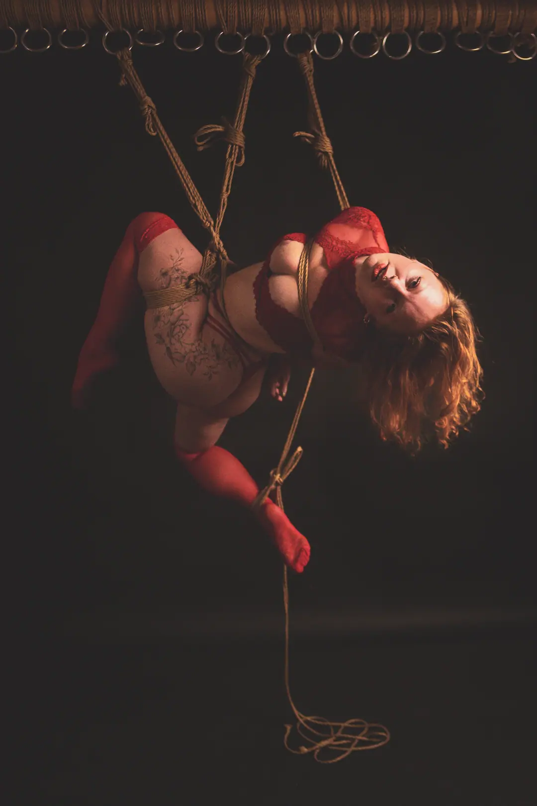Woman wearing red lingerie and socks, suspended in mid-air by intricate rope bondage against a dark background.