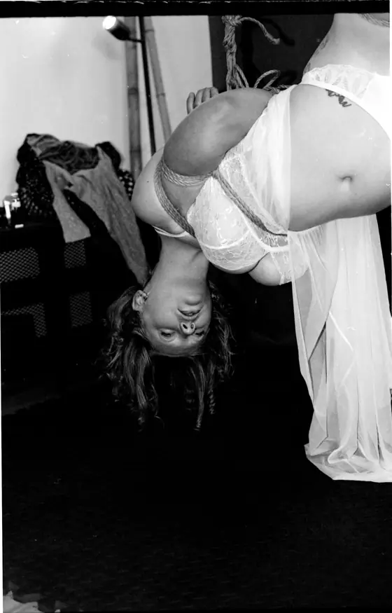 Person wearing white lingerie suspended upside down by rope harness indoors.