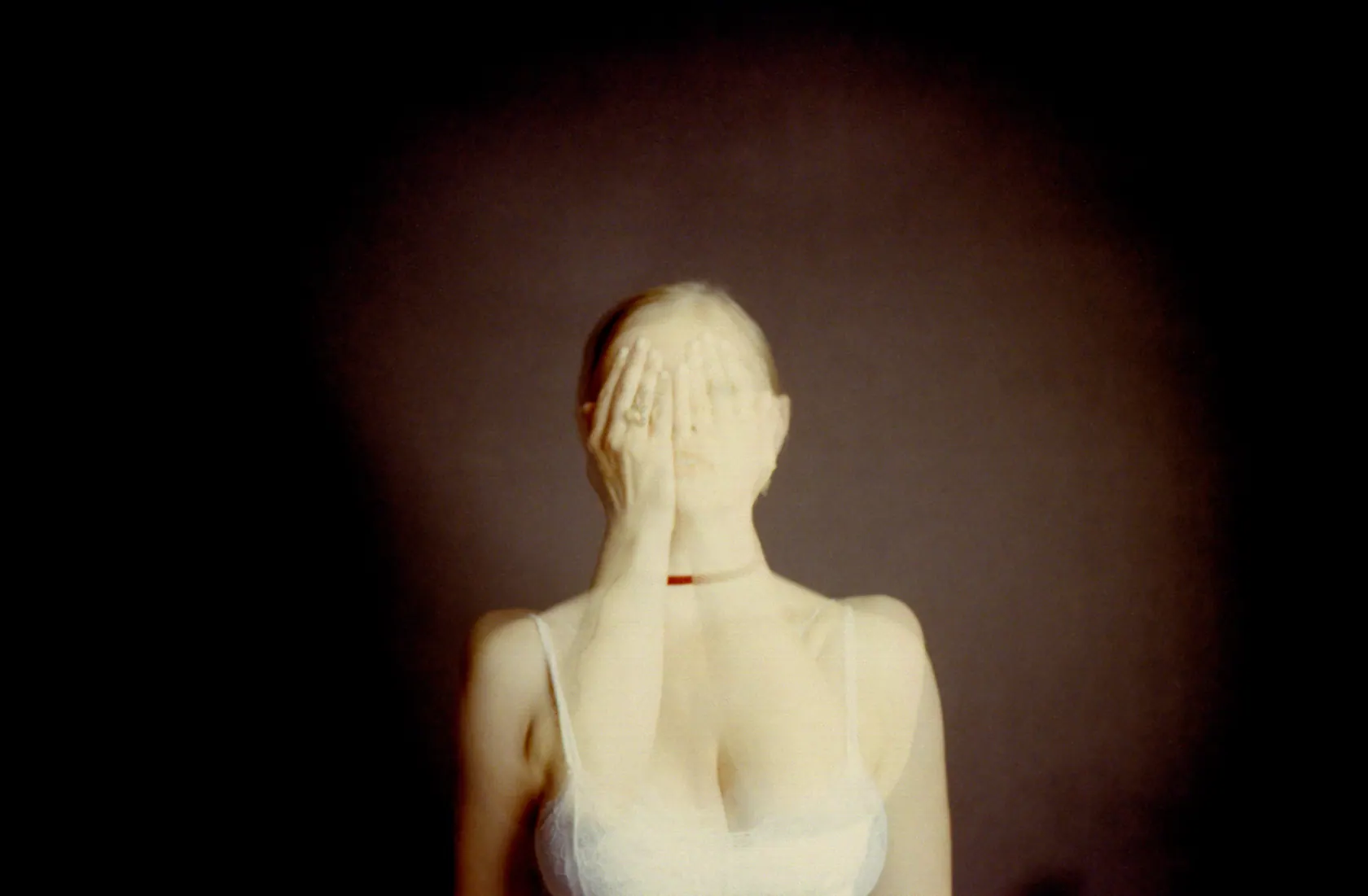 Blurry double exposure of a woman in a white top covering one eye with her hand against a dark background.