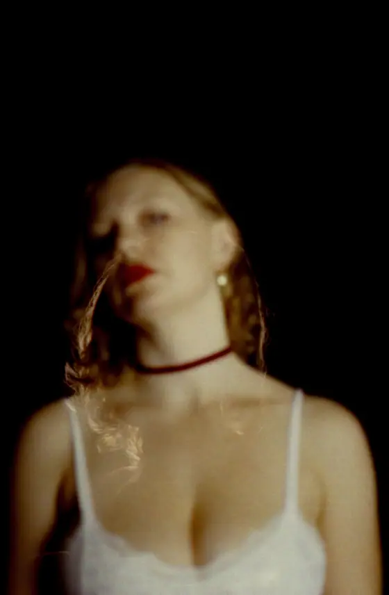 Blurred portrait of a woman with red lipstick, curly hair, wearing a white spaghetti strap top and a choker necklace against a dark