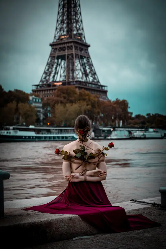 Woman with back exposed and arms tied with rope holding red roses, sitting by a river with the Eiffel Tower and boats in the background.