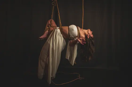 Woman in white flowing skirt suspended in midair with intricate rope harness, dark background, head tilted backwards and hair hanging down.