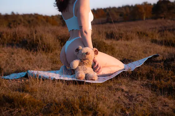 Woman kneeling on a blanket in a field during golden hour, wearing white lingerie and holding a teddy bear behind her back.