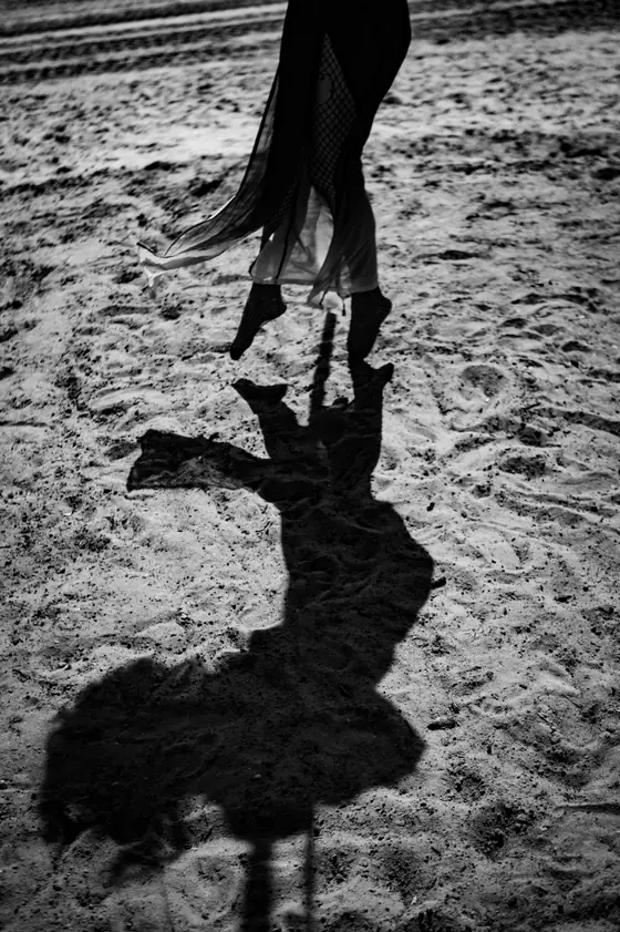 Feet suspended in the air wearing fishnet stockings and a flowing skirt, casting a distinct shadow on textured sandy ground.