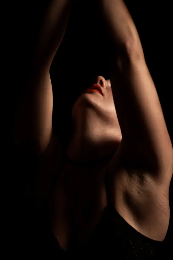 Woman with red lipstick raising arms above head, face partially lit against a dark background.