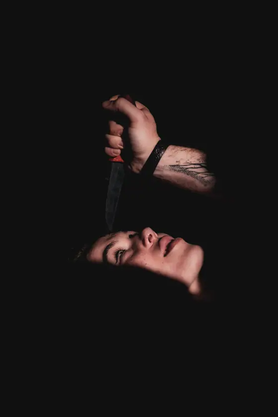 Hand holding a knife above a person's face in a dark setting, with the person's head tilted back and eyes closed.