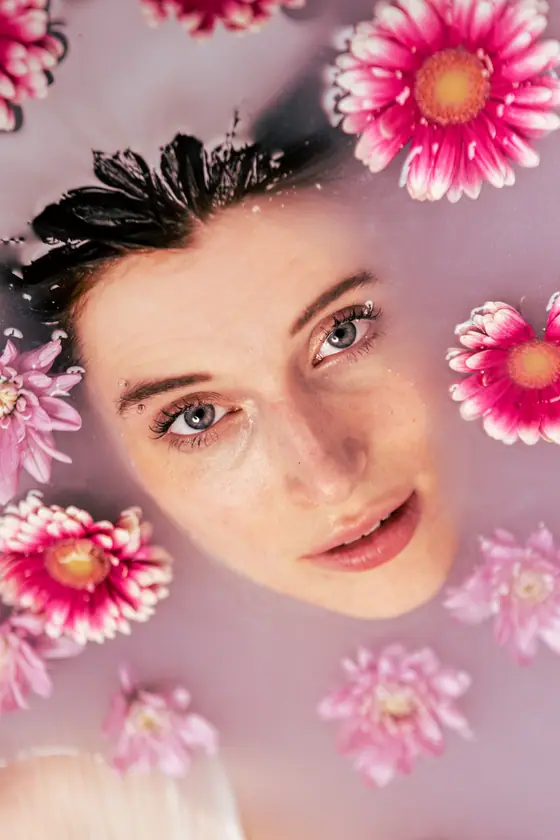 Woman’s face with blue eyes partially submerged in milky water surrounded by pink flowers.