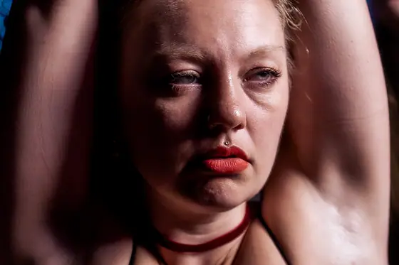 Close-up of a woman with red lipstick and a nose piercing, arms raised behind her head, looking thoughtfully to the side.