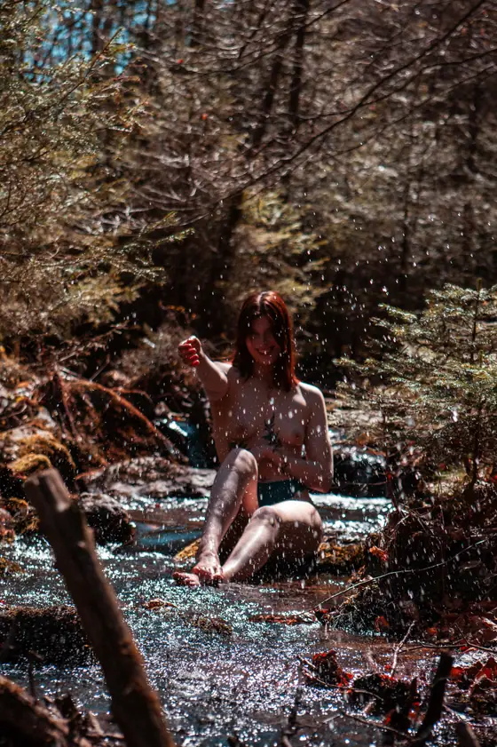 Person sitting in a shallow forest stream, splashing water with their hand, surrounded by trees and natural foliage.