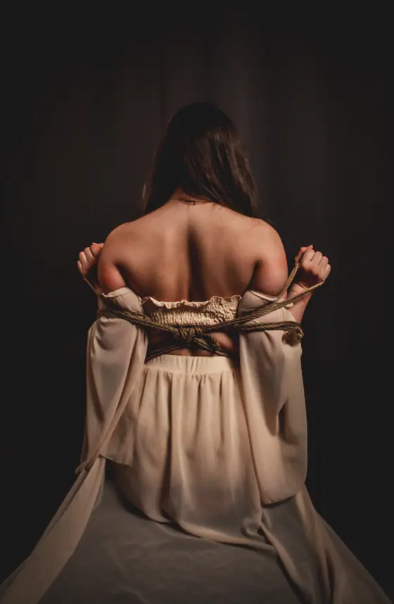 Woman with long hair sitting with her back to the camera, arms tied behind her with rope, wearing a flowing beige dress.