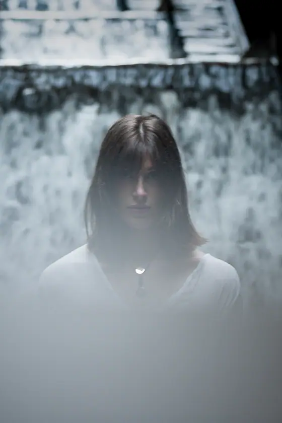 Young woman with shoulder-length hair wearing a white top and necklace, partially obscured by fog, standing in front of a waterfall.