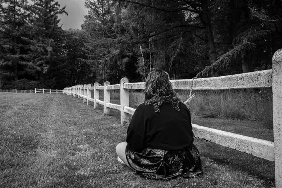 Person with curly hair sitting on grass facing a white fence in a wooded area, black and white photo.