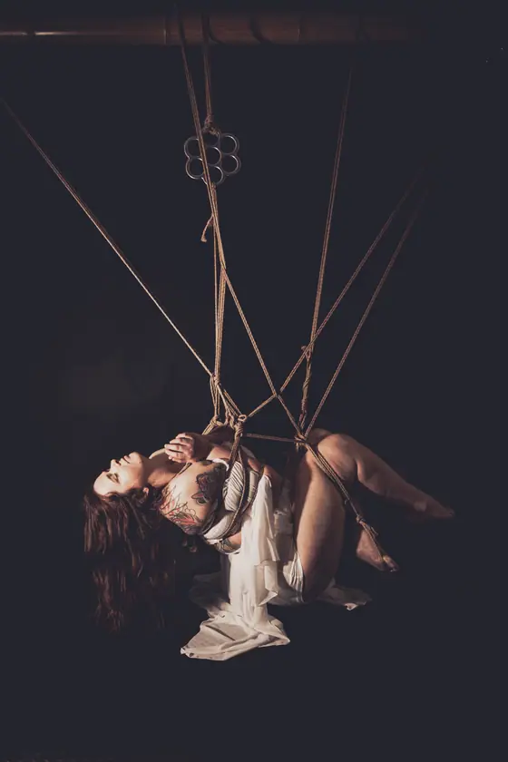 Woman suspended horizontally by multiple ropes in a shibari style against a dark background, wearing white fabric and showing tattoos on