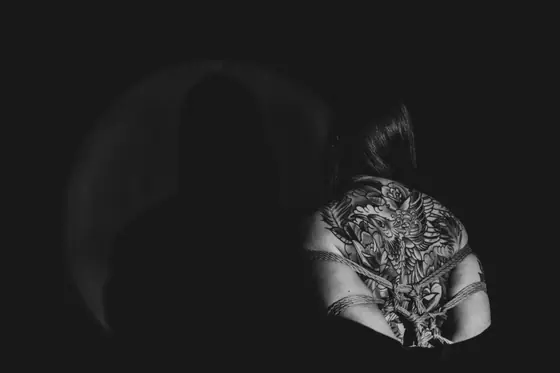 Person with detailed full-back tattoo of a bird, arms tied behind back with rope, sitting against a dark background with a circular light