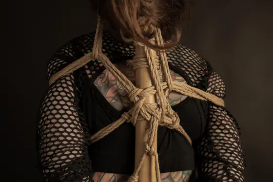 Person wearing a black fishnet top and black bra, with tattoos visible on their back, tied with intricate rope bindings around their torso.