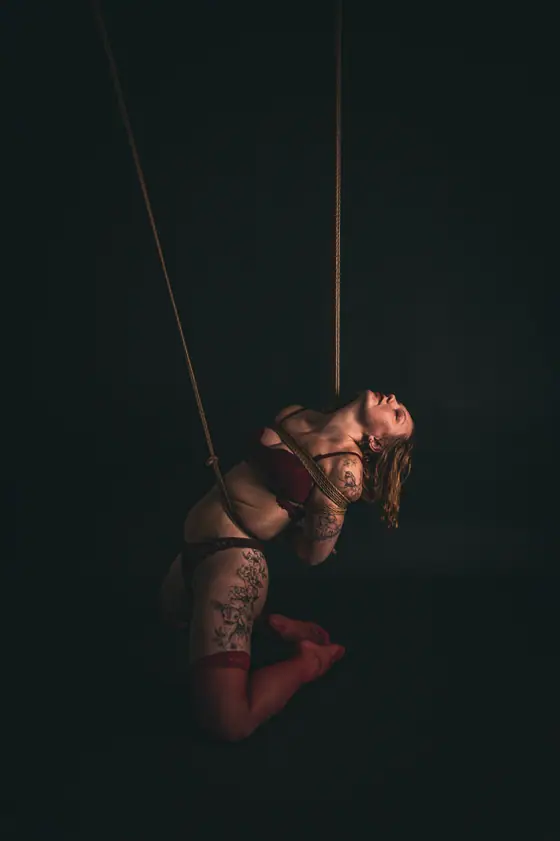 Woman in burgundy lingerie with tattoos, bound with ropes in an artistic pose against a dark background.